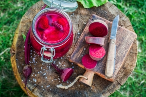 overhead photo of a jar of pickled beets and a cutting board with a fresh beet and knife on it all sitting on a round slab of wood